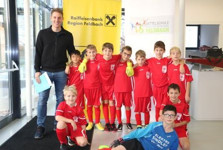 Ein Mann steht hinter einer Gruppe von Jungen in roten Uniformen und lächelt für ein Foto. Sie sind wahrscheinlich Fußballspieler.