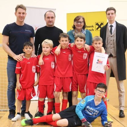 Eine Fußballmannschaft mit jungen Spielern in roten Trikots, begleitet von Trainern, steht vor einem Tor. Ein Junge in einem blauen Shirt hält ein Zertifikat.