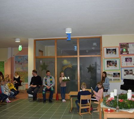 Eine Gruppe von Menschen sitzt und steht in einem Raum. Zwei Erwachsene sitzen auf einem Stuhl vor einem Fenster. Ein Mädchen hält einen Kranz mit brennenden Kerzen. Ein Klavier steht rechts.
