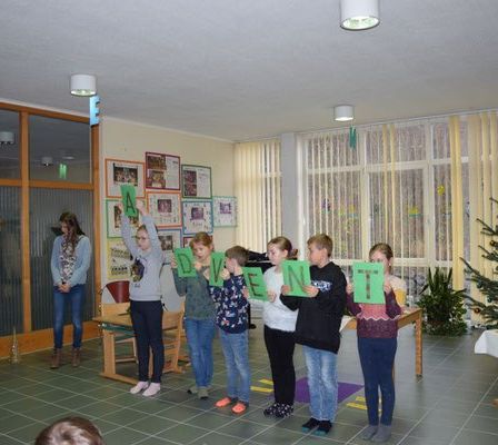 Gruppe von Kindern, die grüne Buchstaben halten und in einem Raum mit Weihnachtsbaum und Dekorationen stehen.