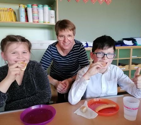 Eine lächelnde Lehrerin mit zwei Schülern in einem Klassenzimmer, einem Mädchen und einem Jungen, essen mit erhobenem Daumen.