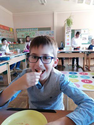 Ein Junge mit Brille sitzt an einem Schreibtisch in einem Klassenzimmer und isst von einem gelben Teller. Im Hintergrund sind andere Schüler und Lehrer zu sehen.