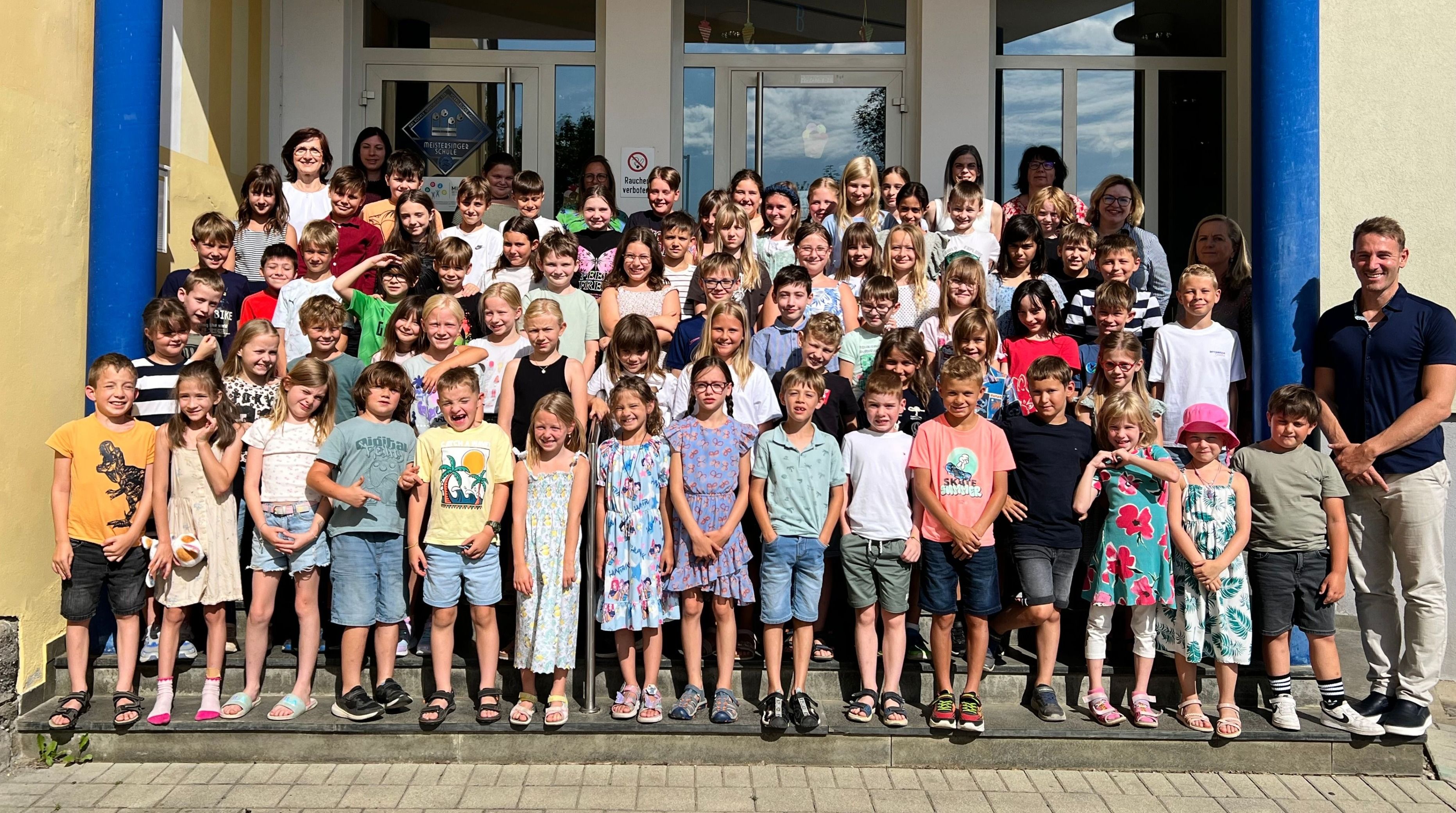 Eine große Gruppe von Kindern und Erwachsenen versammelt sich auf den Stufen eines Gebäudes und lächelt für ein Gruppenfoto. Die Kinder tragen lässige Sommerkleidung. Hinter ihnen steht ein Schild mit der Aufschrift 'Rauchen verboten'.