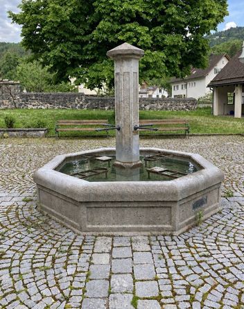 Ein Steinbrunnen mit einer zentralen Säule befindet sich in einem gepflasterten Innenhof, umgeben von Bänken und Grünflächen. Hinter dem Brunnen steht eine Steinmauer und im Hintergrund Häuser.