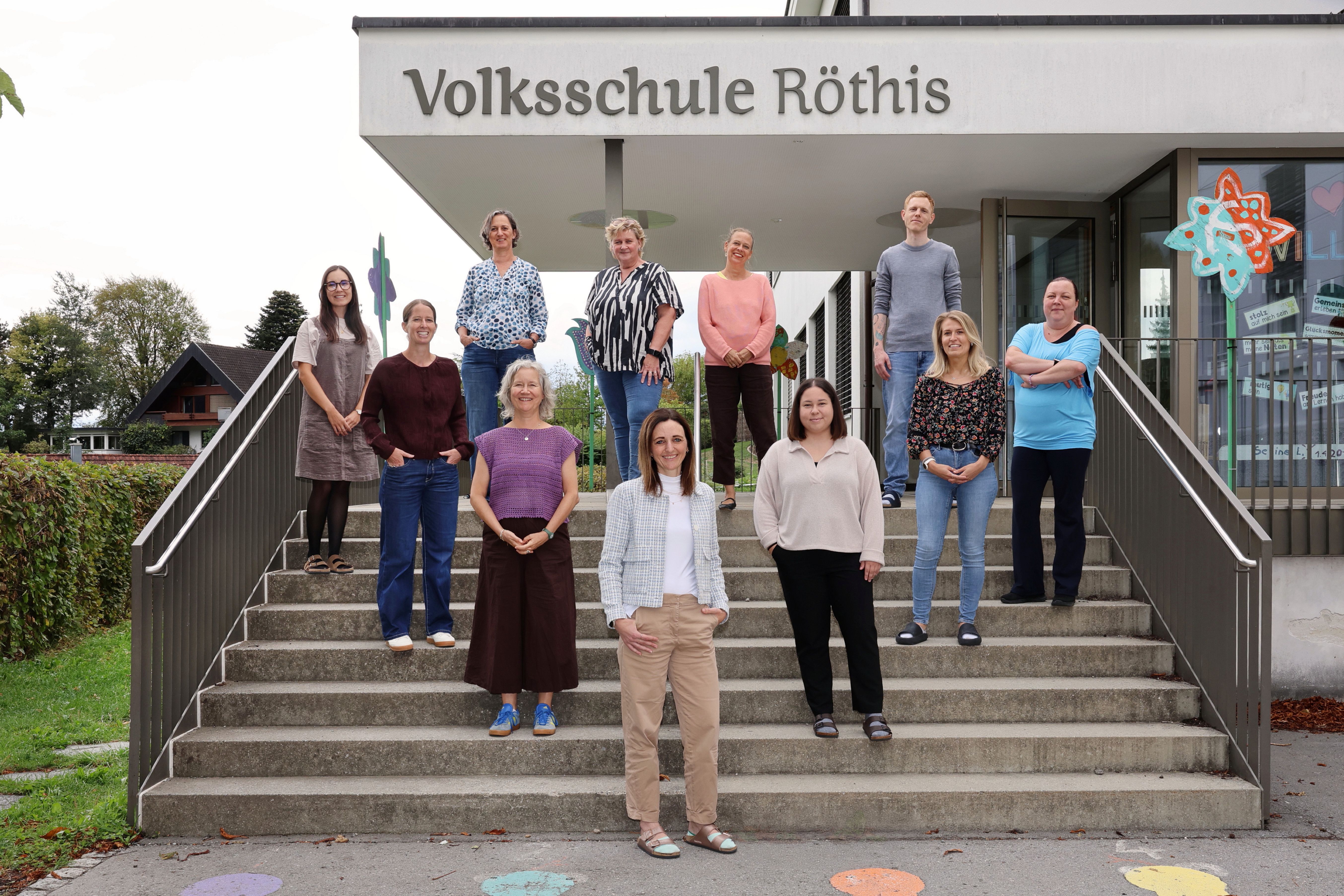 Eine Gruppe von Menschen steht auf den Stufen eines Gebäudes, lächelt und posiert für ein Foto. Das Gebäude hat ein Schild mit der Aufschrift 'Volksschule Rothis'.