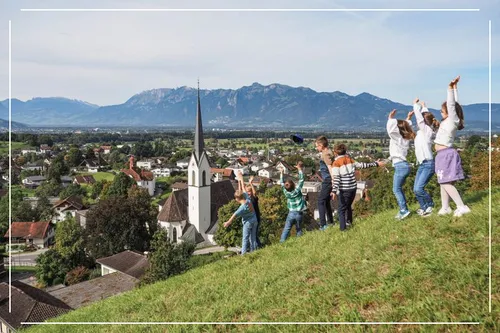Kinder spielen fröhlich auf einem Hügel, mit einer Kirche und einem Dorf darunter, unter einem klaren Himmel mit Bergen im Hintergrund.