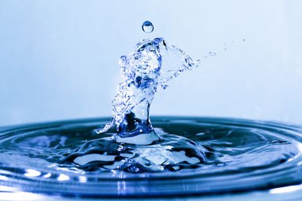 Ein Wassertropfen spritzt in einen Pool und erzeugt Wellen und Tröpfchen. Das Wasser ist klar und blau.