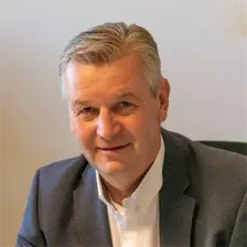 Ein Mann in einem grauen Anzug sitzt und lächelt für ein Foto. Er hat kurze Haare und sitzt auf einem schwarzen Stuhl.
