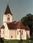 Eine traditionelle weiße Kirche mit einem braunen Dach und einem Turm steht auf einem Rasenfeld, mit Bäumen im Hintergrund.