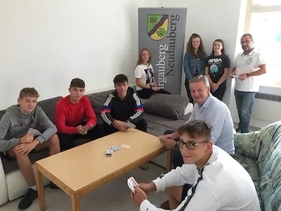 Eine Gruppe von Menschen, sowohl sitzend als auch stehend, versammelt sich um einen Tisch in einem Raum. Einige spielen Karten, andere beobachten. Ein Banner mit dem Text 'Neudauberg' ist an der Wand angebracht.