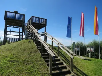 Eine Holztreppe führt zu einem Aussichtsturm auf einem grasbewachsenen Hügel. Drei Fahnen stehen rechts.