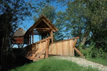 Eine Holzbrücke führt zu einem hölzernen Turm mit einem Strohdach. Die Brücke ist von viel Grün umgeben und der Himmel ist klar und blau.