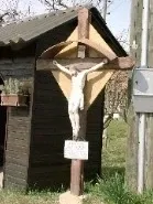Eine kleine Holzstruktur mit einem Kruzifix davor. Das Kruzifix ist hölzern und hat ein Schild darunter. Es gibt eine Topfpflanze an der Seite des Gebäudes.