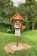 Ein Vogelhaus mit einem Schild darauf steht auf einem Rasen. Das Vogelhaus hat ein Bild einer Person darauf. Bäume umgeben das Gebiet.