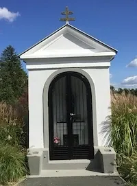 Eine kleine weiße Kapelle mit einer schwarzen Tür und einem Kreuz auf der Spitze. Sie ist von hohem Gras und Bäumen umgeben.