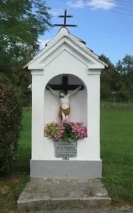 Eine kleine weiße Kapelle mit einem Kreuz oben, mit einer Statue von Jesus Christus am Kreuz und Blumen darunter. Sie steht auf einem Betonsockel in einem Grasfeld.