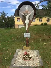 Ein Kruzifix-Gedenkmal steht auf einem grasbewachsenen Hügel vor einem gelben Haus, mit einem kleinen Blumenbeet darunter.