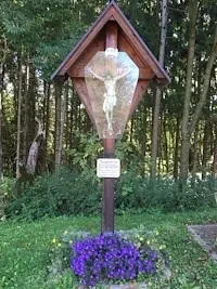 Ein hölzerne Kreuz mit Glasabdeckung und einer Kreuzigungsszene steht in einem Grasbereich mit violetten Blumen und Bäumen im Hintergrund.