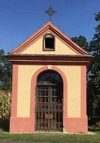 Eine kleine Kapelle mit einem Kreuz oben, in Gelb und Rosa gestrichen, hat einen bogenförmigen Eingang mit einem Metalltor und einem kleinen Fenster darüber.