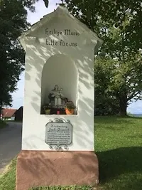 Eine weiße Kapelle mit einer Statue einer Frau im Inneren, umgeben von Pflanzen und Blumen, steht auf einem Rasenfeld. Die Kapelle hat eine Inschrift und befindet sich neben einer Straße.