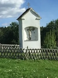 Eine kleine weiße Kapelle mit einem Kreuz auf der Spitze und einem Kruzifix im Inneren. Die Kapelle ist von einem Holzzaun umgeben und befindet sich auf einer Wiese.