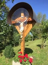 Ein hölzerne Kreuz mit Glasüberzug steht auf einem grünen Feld und zeigt eine gekreuzigte Figur. Es ist von roten Blumen und üppiger Vegetation umgeben.