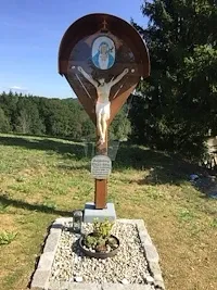 Ein Kruzifix auf einer Holztafel steht in einem Grasbereich, mit einem Topfpflanzen an der Basis und Bäumen im Hintergrund.
