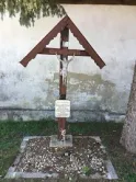 Ein hölzerne Kreuz mit einem Kruzifix steht vor einer verwitterten Wand, in der Nähe eines Baumes. Eine Plakette ist an der Basis des Kreuzes befestigt.