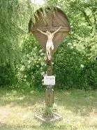 Ein Kruzifix ist an einer Holzstruktur montiert, mit einem kleinen Schild davor, in einem Grasbereich aufgestellt, umgeben von grünen Pflanzen und Bäumen.