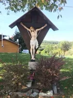 Ein Kruzifix auf einem dreieckigen Holzgestell steht auf einem Rasenfeld mit einem Haus und Pflanzen im Hintergrund.