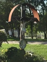 Ein großes Kruzifix befindet sich in einem Rasenbereich, mit Jesus am Kreuz. Das Kreuz steht auf einer runden Basis, und es gibt Bäume und Büsche in der Umgebung.