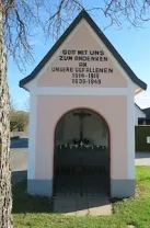 Eine kleine pinke und graue Kapelle mit schwarzem Dach. Der Text lautet 'Gott mit uns zum Andenken an unsere Gefallenen 1914-1918 1939-1945'. Die Kapelle hat einen gewölbten Eingang mit einem Kreuz darüber.
