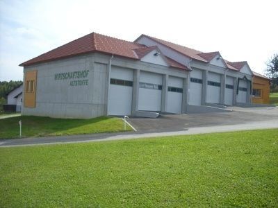 Ein großes graues Gebäude mit rotem Dach und mehreren weißen Garagen ist mit 'Wirtschaftshof Alzstoffte' beschriftet. Es hat eine Einfahrt und ist von einem Grasfeld umgeben.
