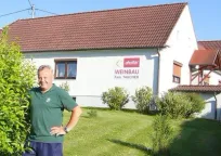 Ein Mann in einem grünen Shirt steht vor einem weißen Haus mit einem roten Dach. Das Haus hat das Schild 'Weinbau Fam. Taucher'. Er hat die Hände in den Hüften.