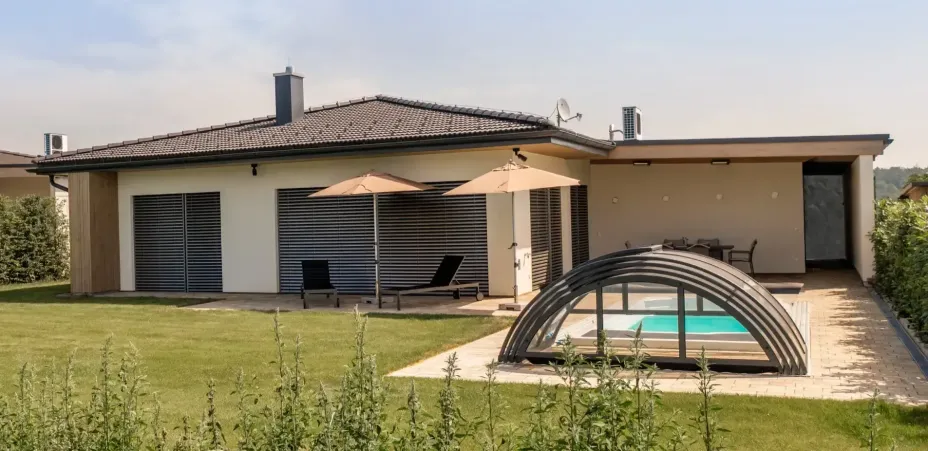 Ein modernes Haus mit einem Schornstein und einer Satellitenschüssel auf dem Dach. Draußen gibt es zwei Liegestühle unter Sonnenschirmen und einen Pool mit Glasüberdachung.