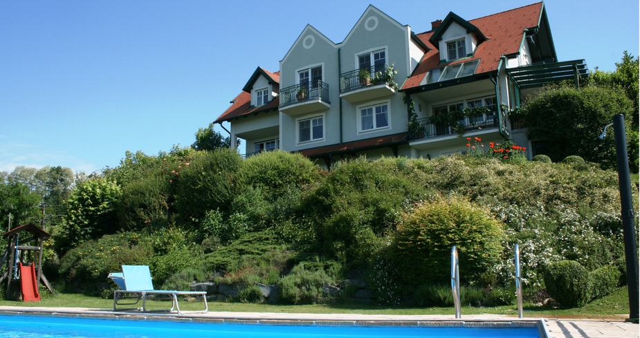 Ein Haus mit roten Dächern und Balkonen steht auf einem Hügel. Davor befindet sich ein Schwimmbad mit einem Stuhl in der Nähe.