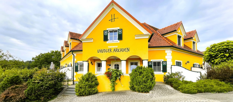 Ein hellgelbes Gebäude mit grünen Fensterläden und dem Schild 'Vhudler Arkaden'. Es hat ein rotes Dach, weiße Verkleidung und Büsche davor.