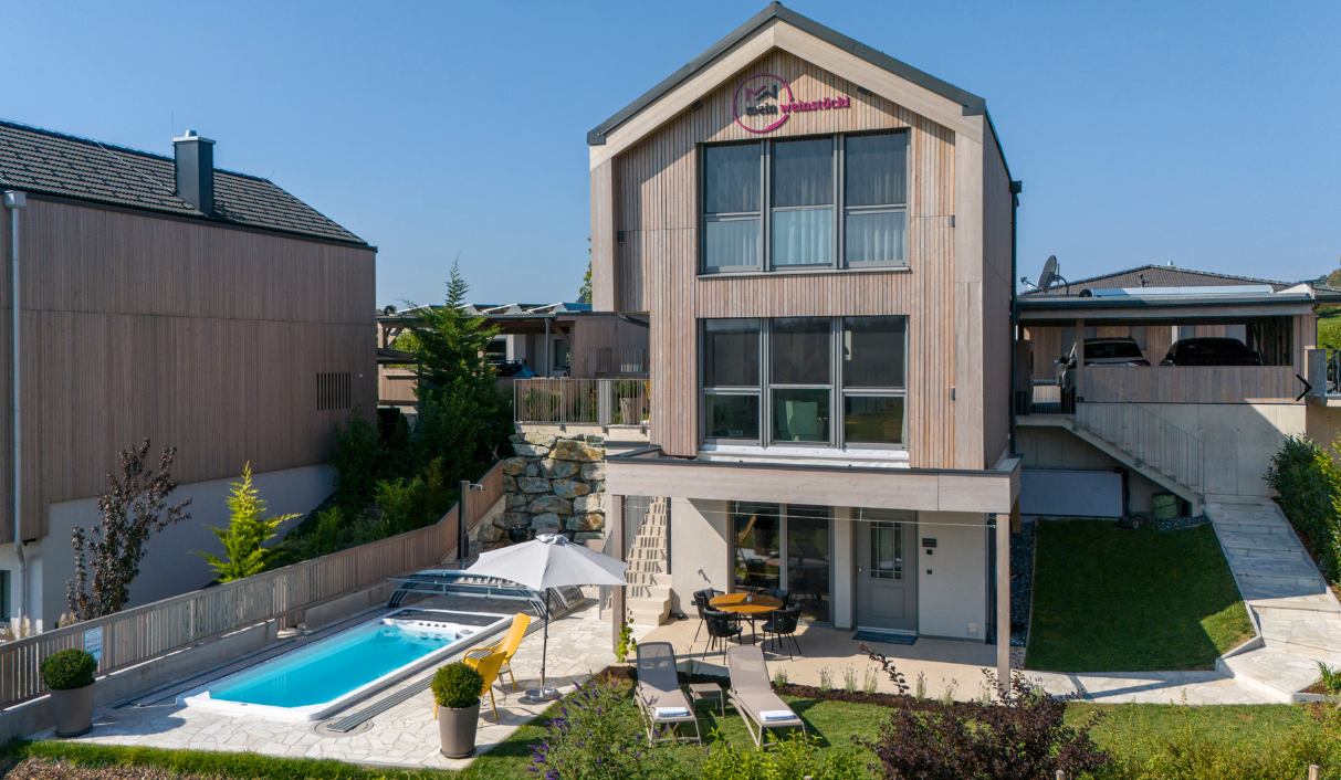 Ein modernes architektonisches Design eines Hauses mit Schwimmbad und Gartenmöbeln. Das Haus hat Glasfenster und ein hölzerne Außenseite.