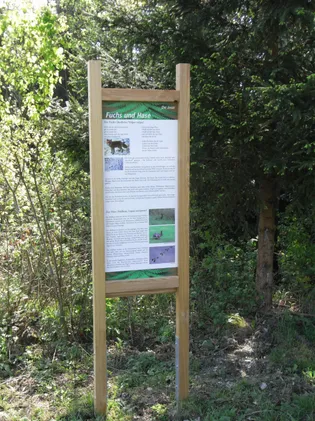 Ein Holzschild mit Text und Bildern von Fuchs und Hase steht in einem Wald. Das Schild trägt den Titel 'Fuchs und Hase' und bietet Informationen über die Tiere.