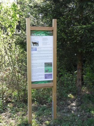 Ein Holzschild mit Text und Bildern von Fuchs und Hase steht in einem Wald. Das Schild trägt den Titel 'Fuchs und Hase' und bietet Informationen über die Tiere.