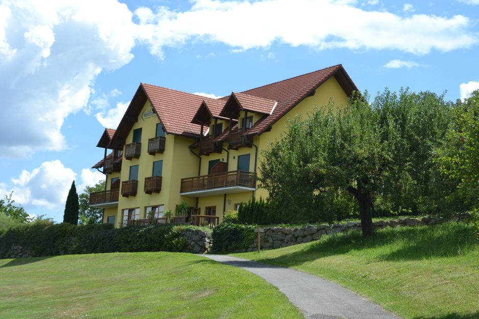 Ein gelbes Gebäude mit einem braunen Dach und mehreren Balkonen steht auf einem grünen Hügel unter einem blauen Himmel mit Wolken.