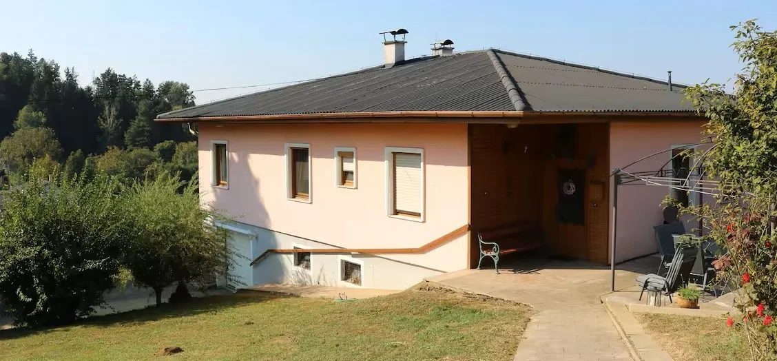 Ein rosafarbenes Haus mit grauem Dach und Schornsteinen, umgeben von Bäumen und Gras. Vor dem Haus steht eine Bank.