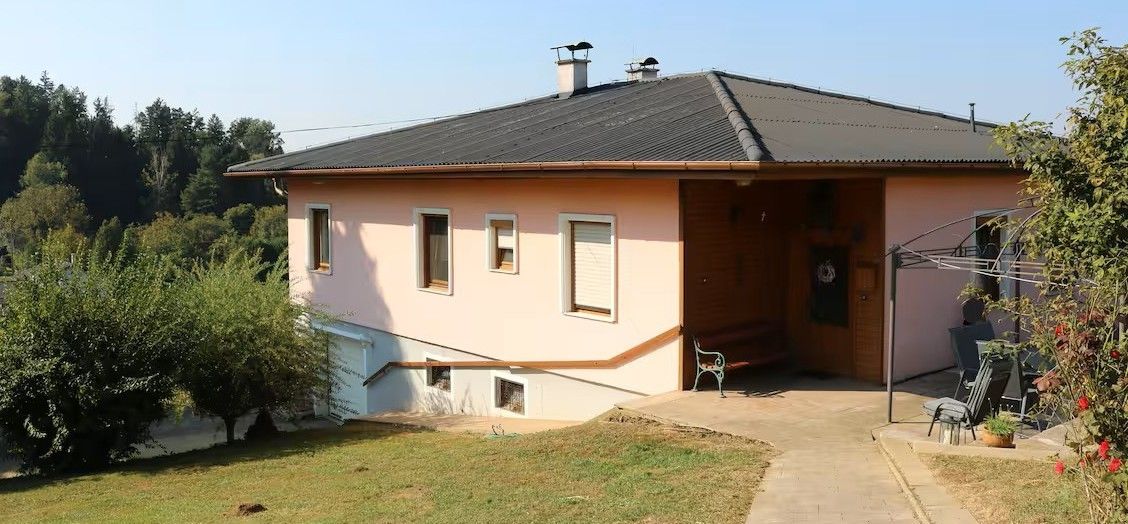 Ein rosafarbenes Haus mit grauem Dach und Schornsteinen, umgeben von Bäumen und Gras. Vor dem Haus steht eine Bank.