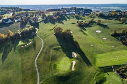 Luftaufnahme eines Golfplatzes mit einem gewundenen Pfad, mehreren Löchern und einem Haus in der Ferne. Der Platz ist von Bäumen und Grünflächen umgeben.