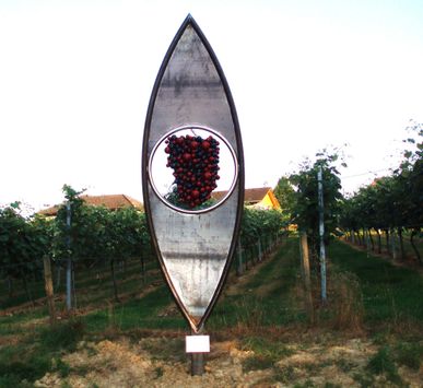 Eine Metallskulptur in Form eines Blattes mit einem kreisförmigen Ausschnitt in der Mitte, der eine Ansammlung roter Beeren zeigt. Die Skulptur befindet sich in einem Weinberg mit Rebenreihen und einem Haus im Hintergrund.