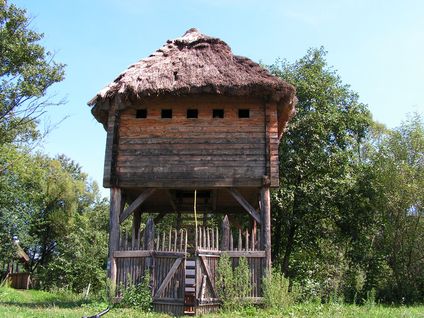 Ein alter Holzturm mit Strohdach und Holzzaun steht auf einem Grasfeld mit Bäumen im Hintergrund.
