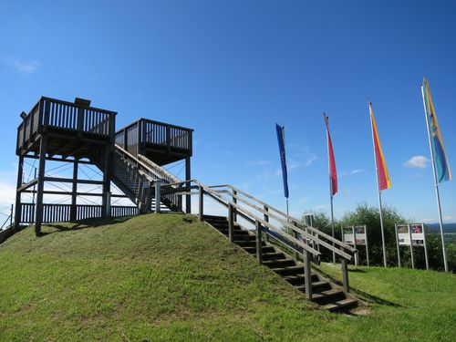 Eine hölzerne Treppe führt zu einer hölzernen Plattform mit einem Geländer. Drei Fahnen stehen auf Stangen auf einem Grashügel. Der Himmel ist blau mit einigen Wolken.