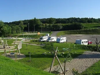 Ein Luftbild eines Campingplatzes mit auf Gras geparkten Wohnmobilen, einem Spielplatz und einem roten Auto auf der Straße. Bäume und Straßenlaternen umgeben das Gebiet.
