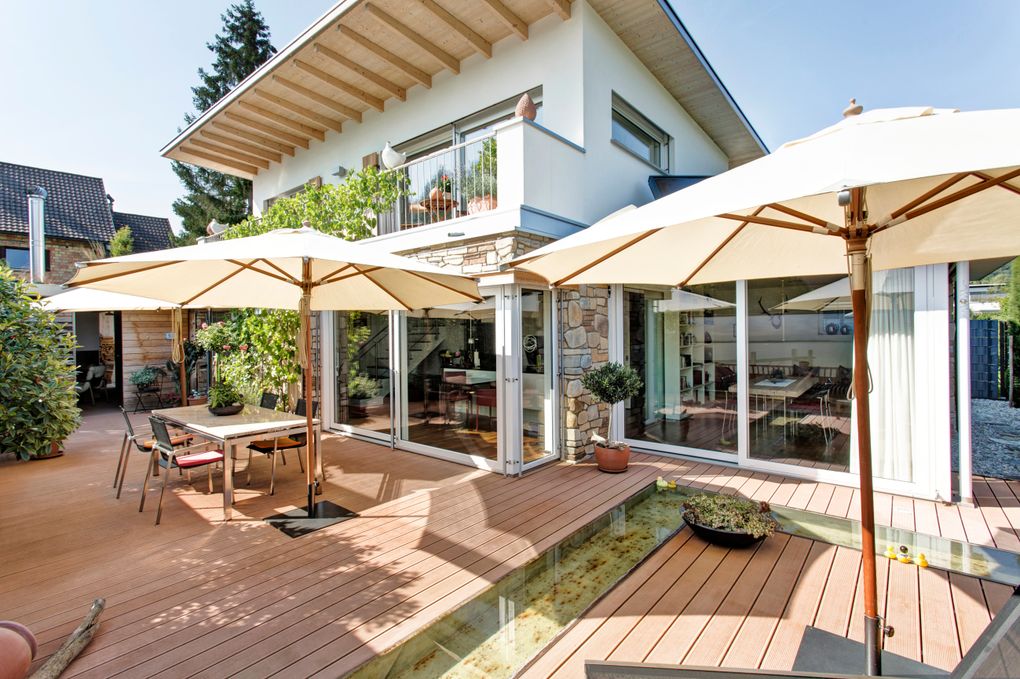  Abschnitt Bild: Ein modernes Haus mit Glastüren und -fenstern, einem Balkon mit Pflanzen und einer Terrasse mit Tischen, Stühlen und Sonnenschirmen. Es gibt ein kleines Wasserspiel und Topfpflanzen auf der Terrasse.