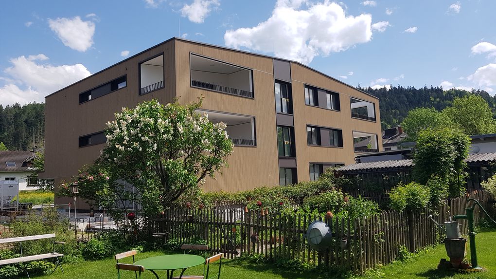  Abschnitt Bild: Ein modernes Mehrfamilienhaus mit Balkonen und Fenstern ist von einem Holzzaun und einem Garten mit einem grünen Tisch und Stühlen umgeben.